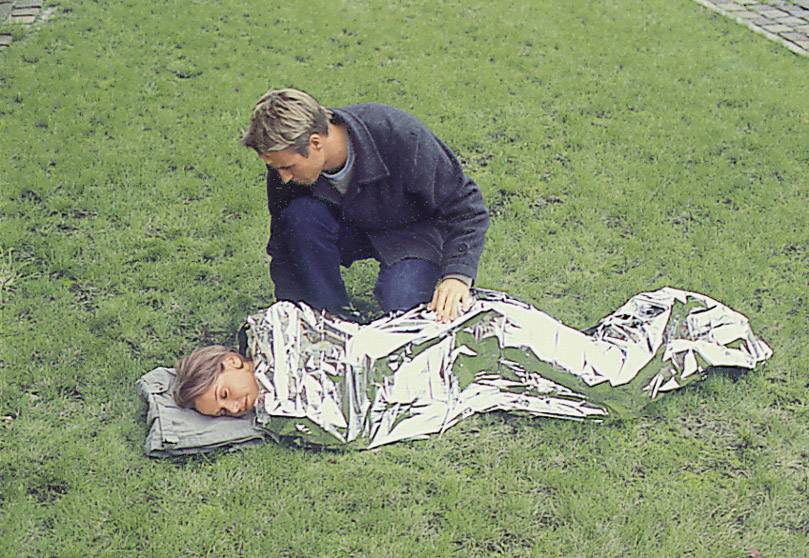 A person is lying wrapped in a thermal blanket on the grass, while another person leans over them anxiously.