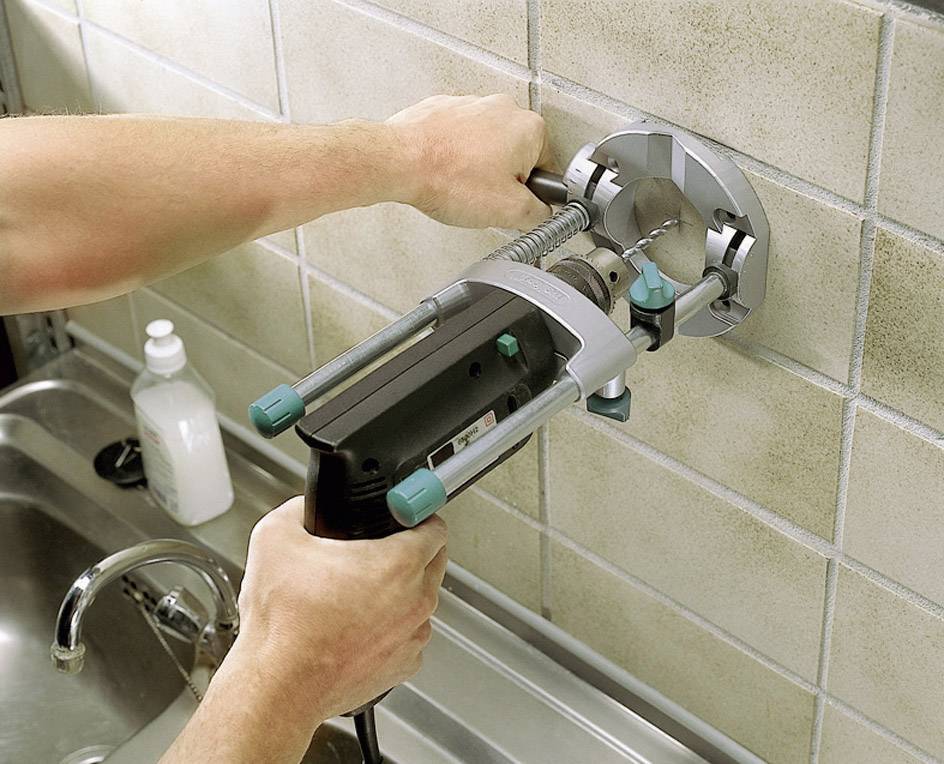 A person is working with an electric drill on a tiled wall above a washbasin.