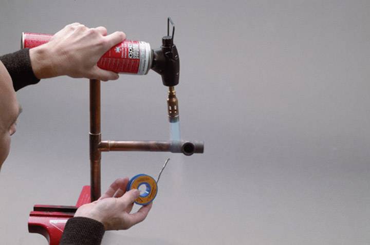 A person is soldering two copper pipes together while holding a blowtorch. One pipe is secured in a vice.