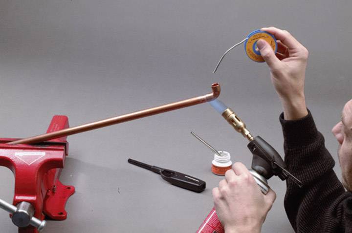 A person is soldering a copper pipe that is secured in a red vice. They are holding a blowtorch in their right hand and solder in their left hand.