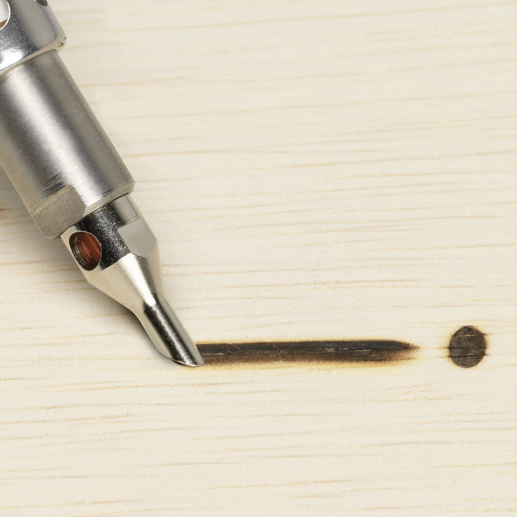 A soldering iron with a fine tip burns a narrow line and a point into a light-coloured wooden board.