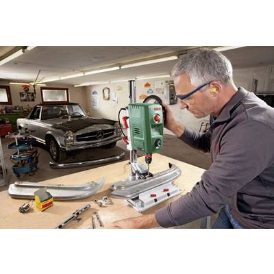 A man is working on a drill press in a workshop, with a classic car standing in the background.