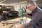 A man is working on a drill press in a workshop, with a classic car standing in the background.