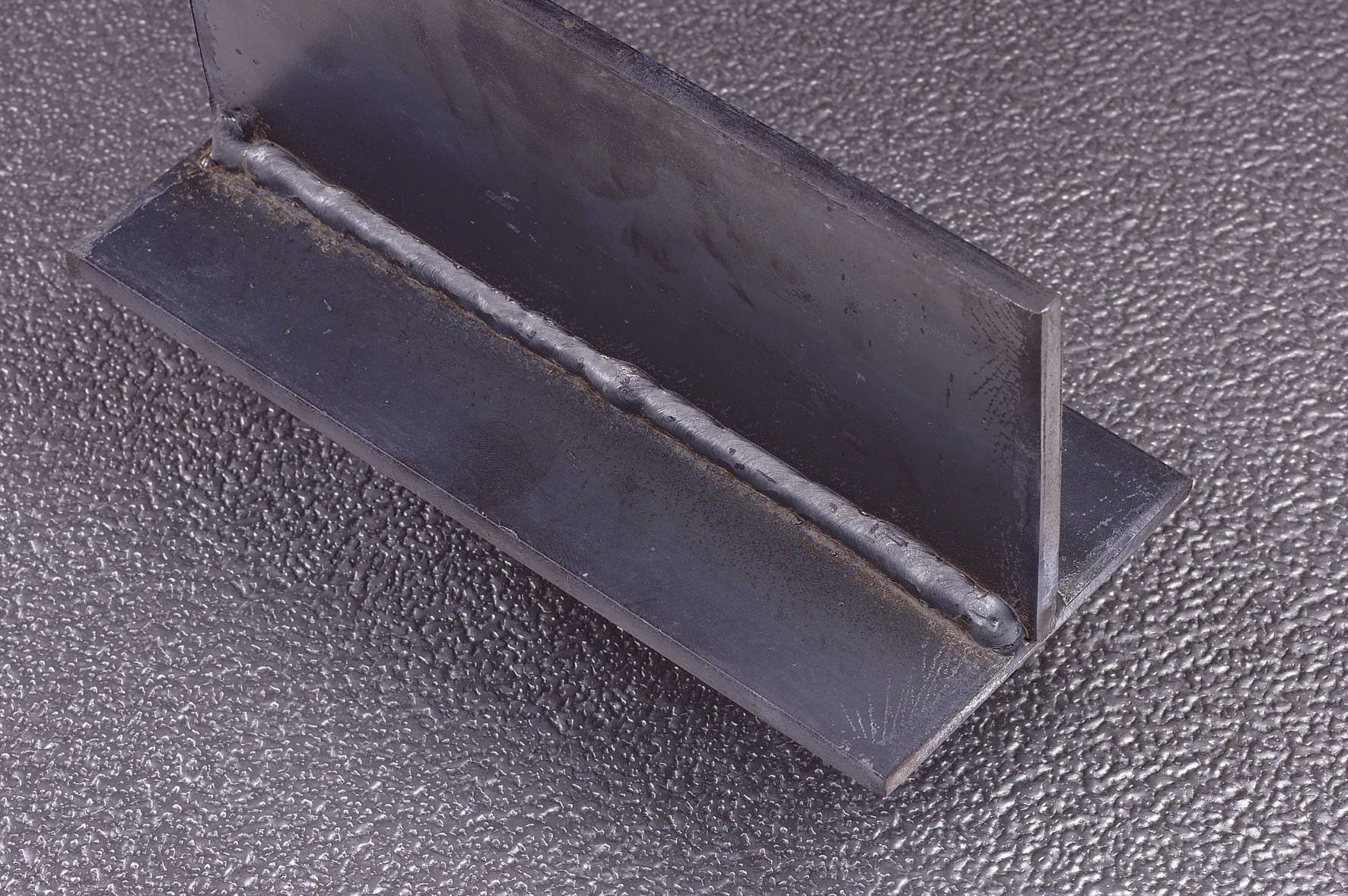 A weld joint in a metal connection, arranged in an L-shape, on a structured, grey surface.