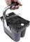 A hand is holding a black battery carrier with a handle. The battery has plus and minus markings on its top surface.