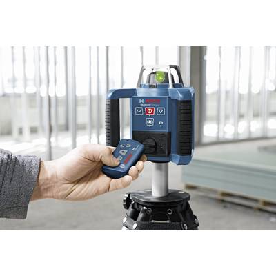 A hand is holding a remote control pointed at a blue laser level device in an unfinished room.