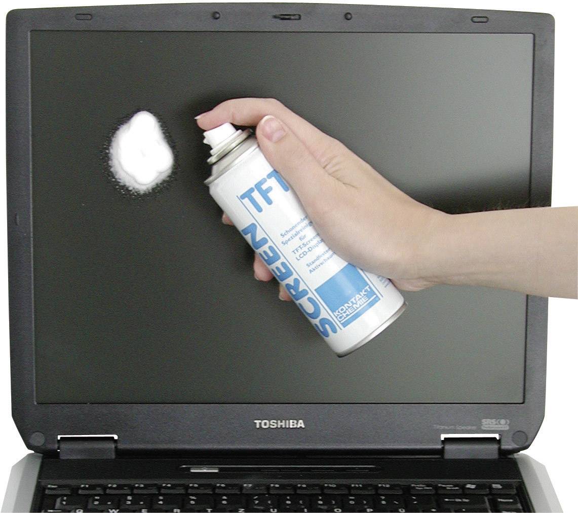 A hand sprays cleaner onto a laptop screen. The screen displays a white foam spot from the spray bottle.