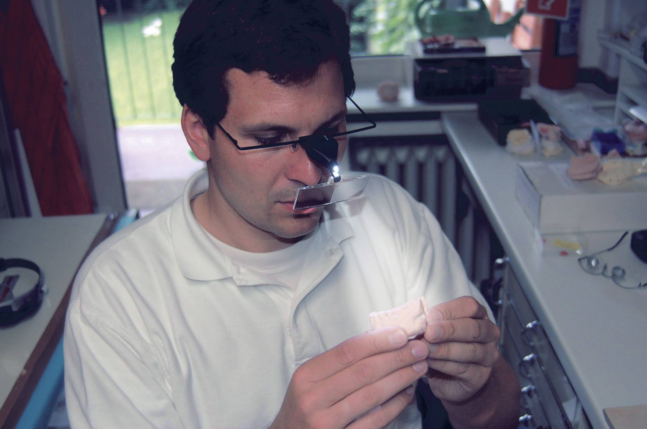 A person in a laboratory is examining a dental prosthesis model using a magnifying loupe. Work equipment is visible in the background.
