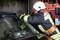 A firefighter in protective gear breaks the windscreen of a car with an axe. A fire engine is parked in the background.