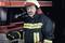 A firefighter in full uniform stands in front of a fire engine, holding a radio handset.