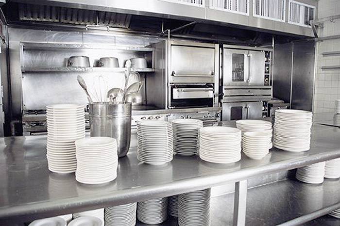 A commercial kitchen environment with stacked plates on a stainless steel worktop in front of large kitchen appliances in the background.