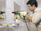 A man wearing safety glasses and gloves is drilling a hole in a bathroom tile above a sink. Tools are laid out ready.