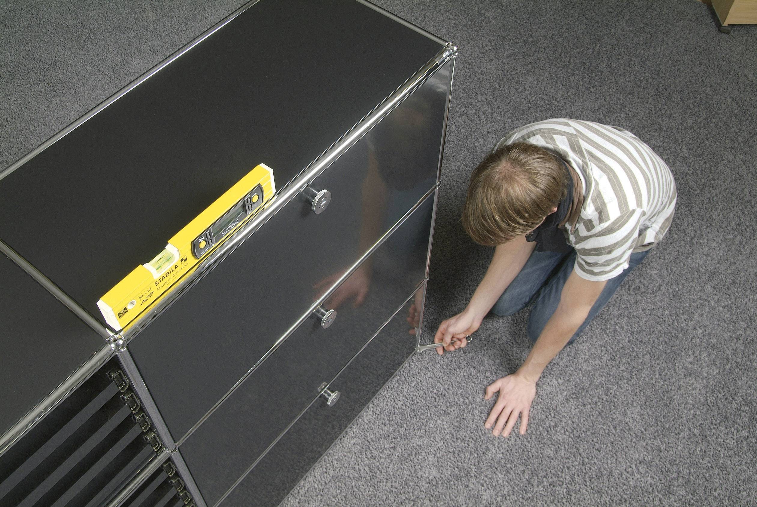 A person is kneeling on the carpet and assembling furniture with an Allen key. A tool is lying on top of the piece of furniture.