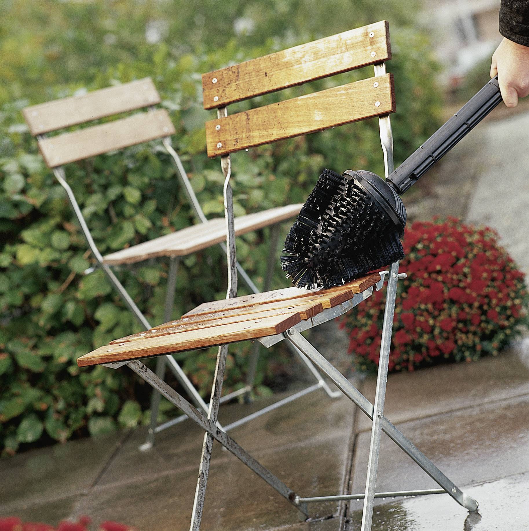 A hand is cleaning a wet, wooden folding chair with a large brush. In the blurry background, a second chair and some bushes are visible.