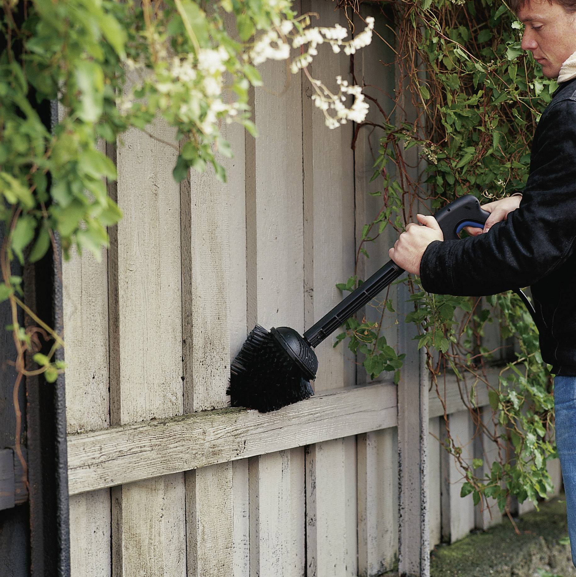 A man is cleaning a wooden wall with a large brush, surrounded by green plants; focus on the cleaning activity.