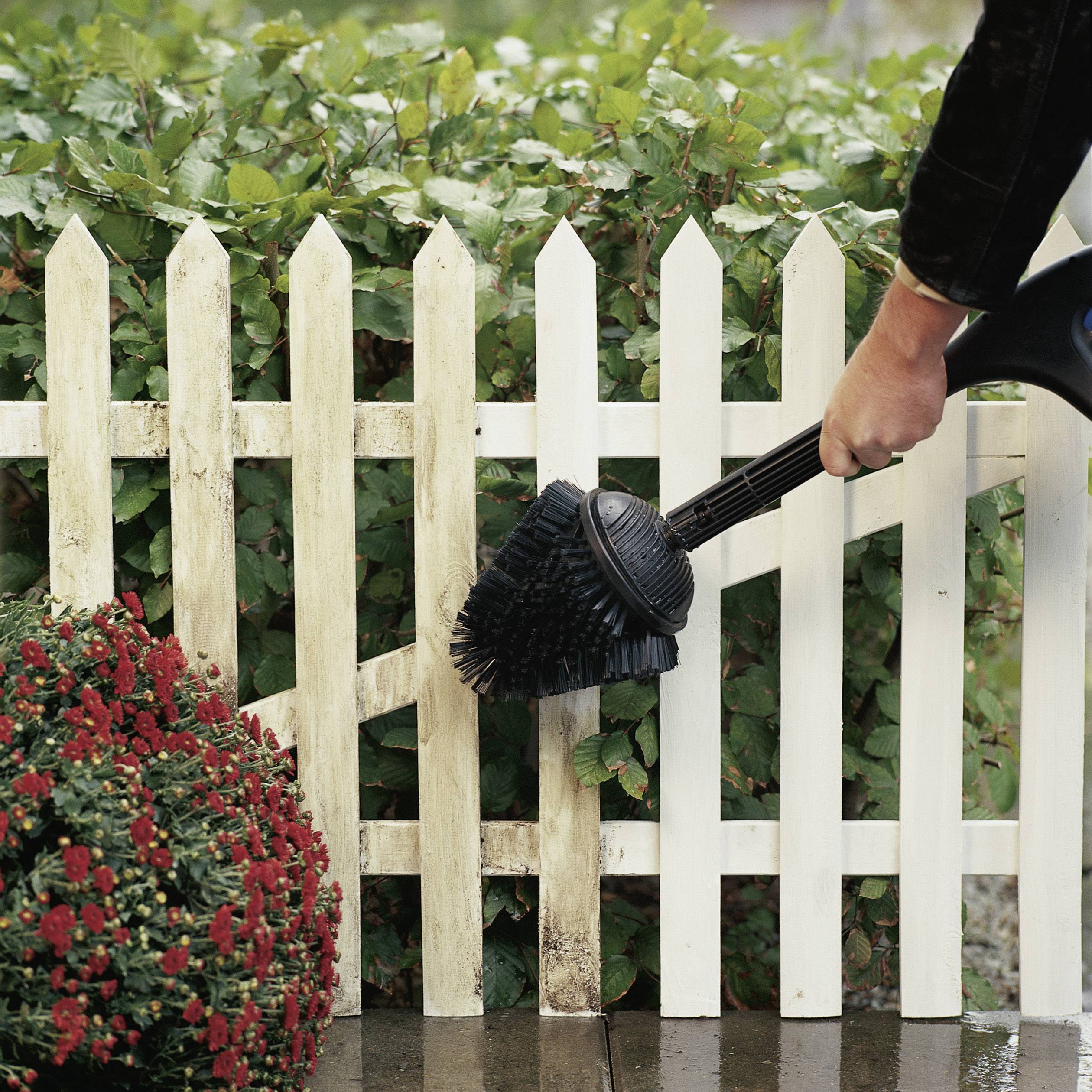 A broom-like tool is held by a hand and cleans a narrow gap between a wooden fence and bushes.