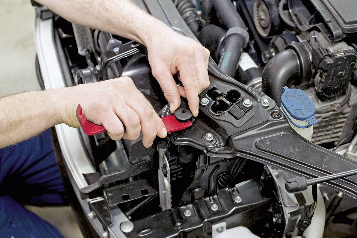 A person opens the bonnet of a car with a red lever handle to access the engine compartment.