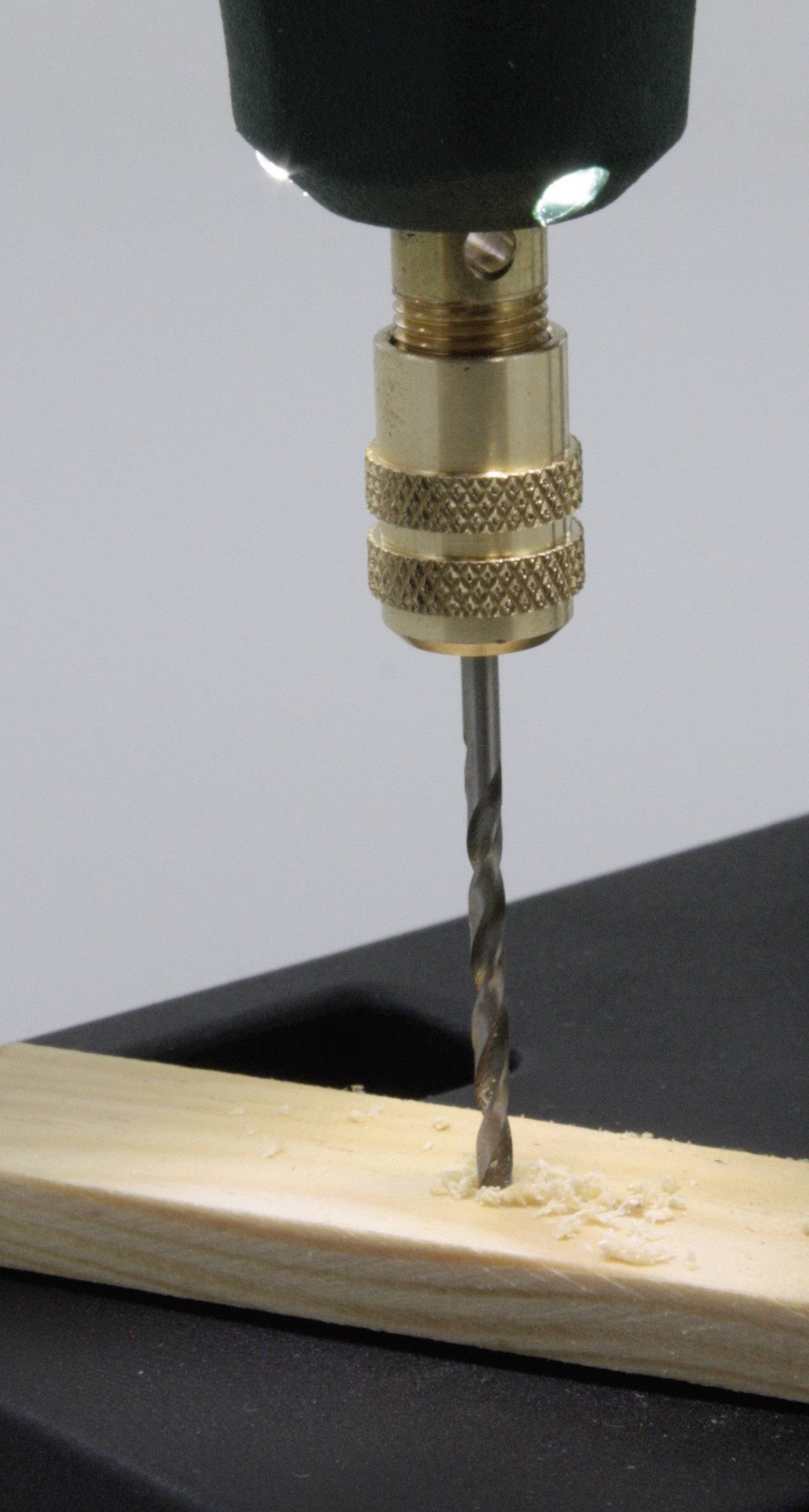 An electric drill is boring through a wooden board. Wood shavings are scattered around the drill hole.