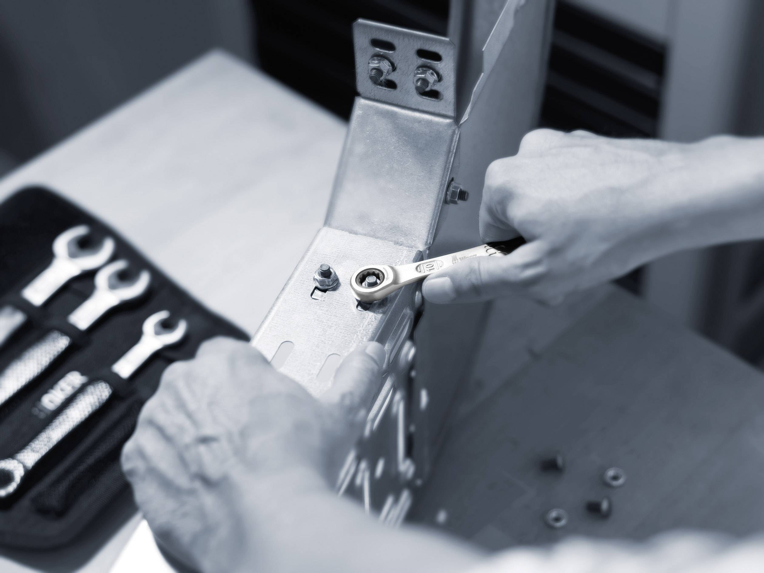 A person is tightening a screw on a metal construction with a spanner. An open toolkit lies in the background.