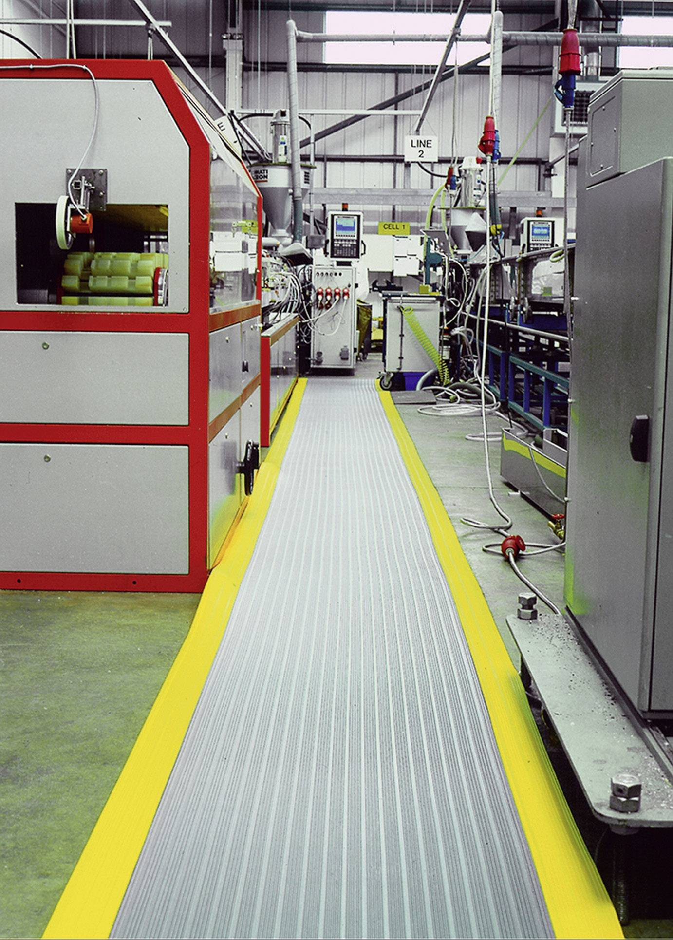 Production line in a factory, machines on both sides of a marked pathway, surrounded by technical equipment and cables.