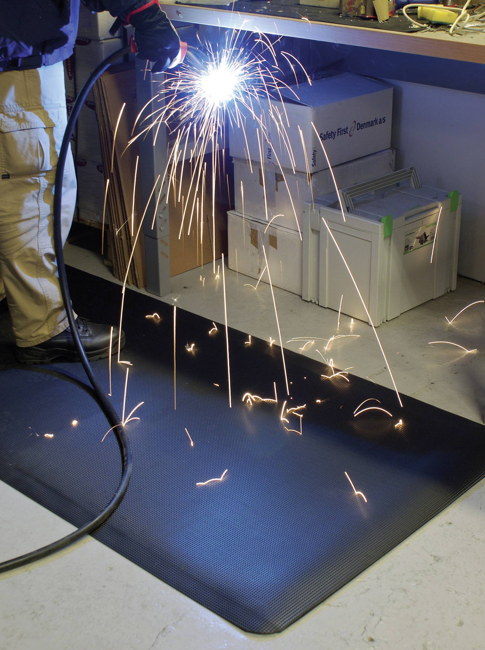 A worker is welding with a handheld welding device, sparks flying on a non-slip floor covering designed for safety.