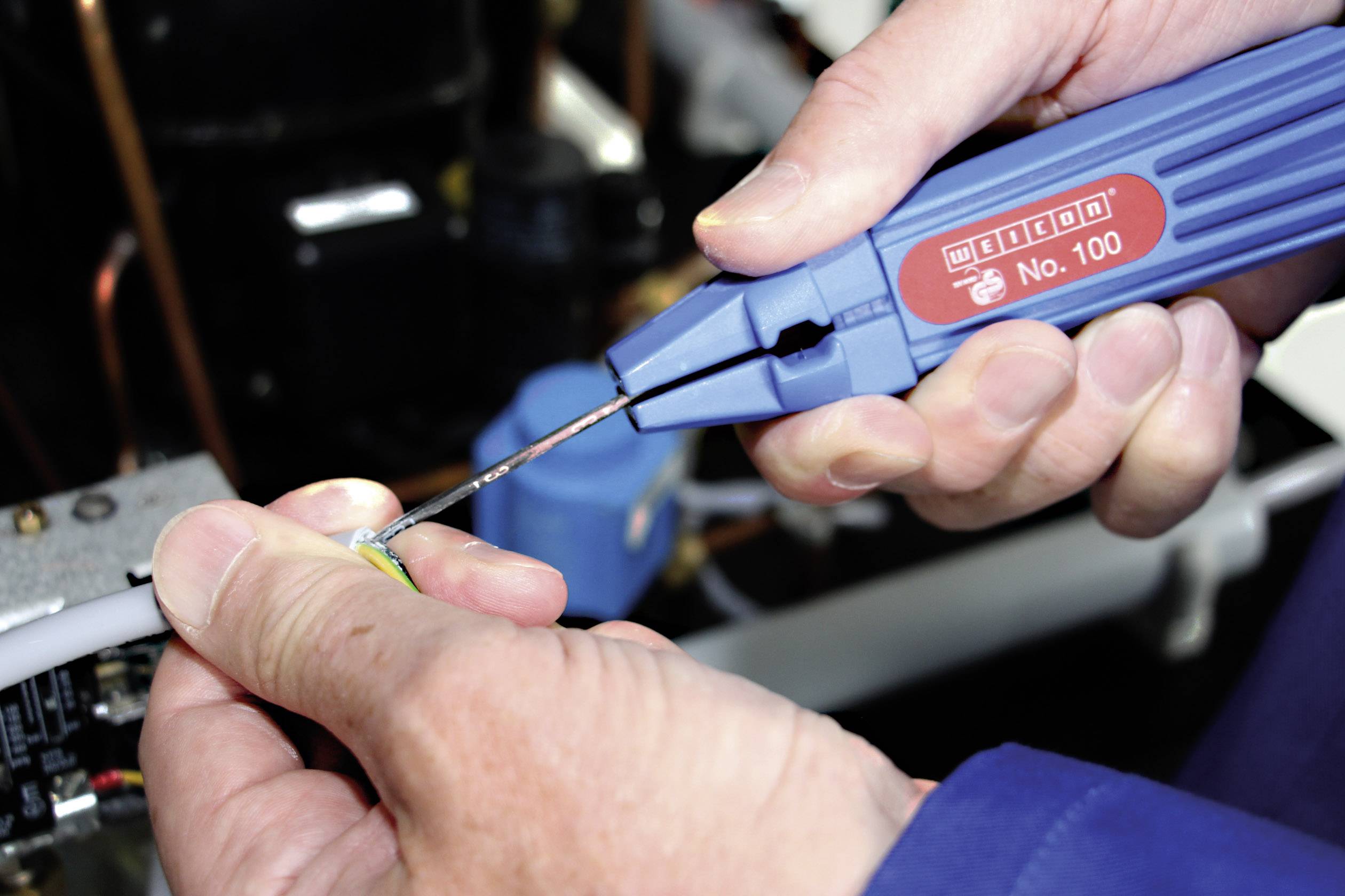 Close-up of hands working on an electrical cable with a blue wire stripping tool. Background blurred with copper wires.