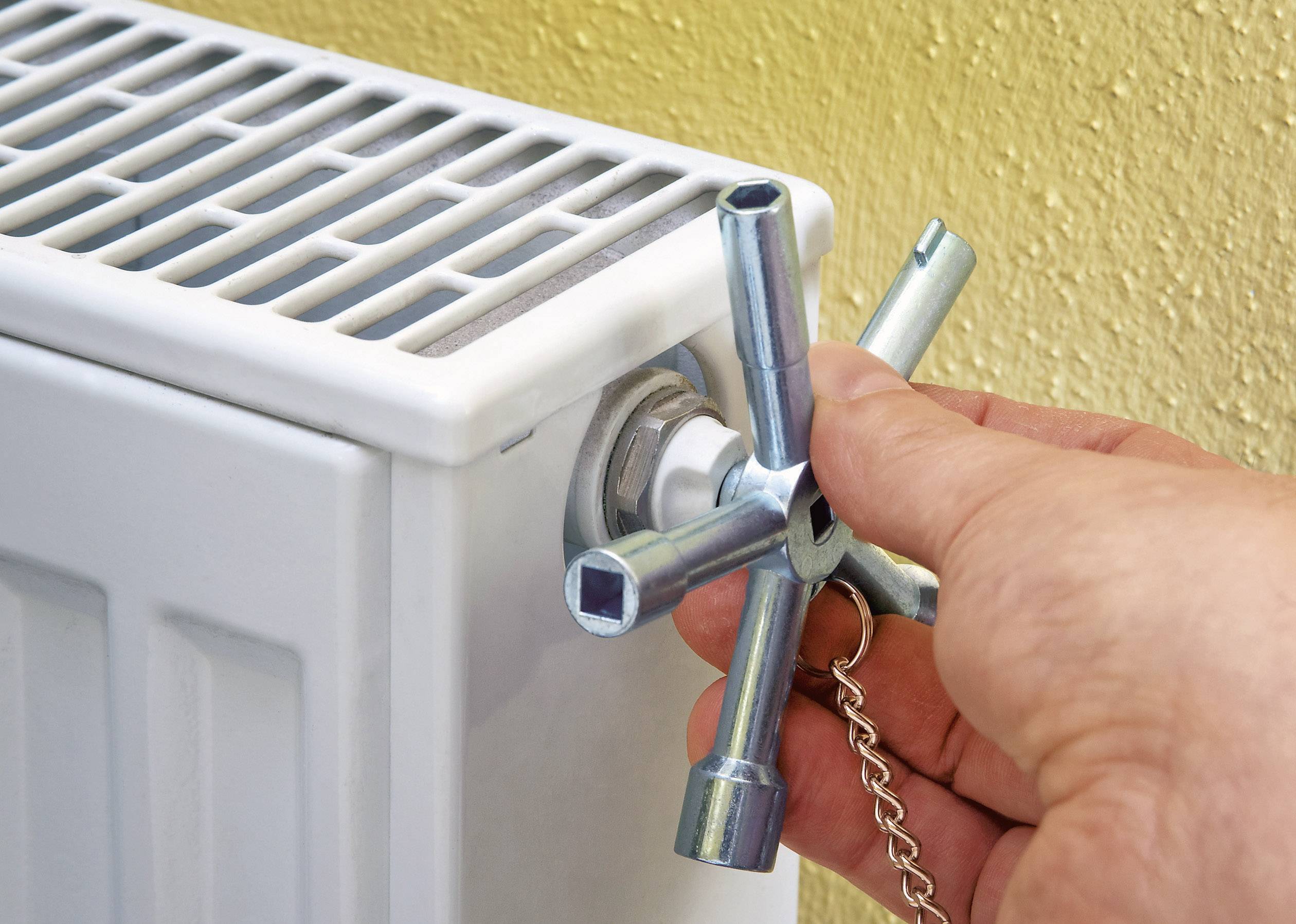 Hand with a spanner turning a radiator valve to bleed the heating. Yellow wall in the background.