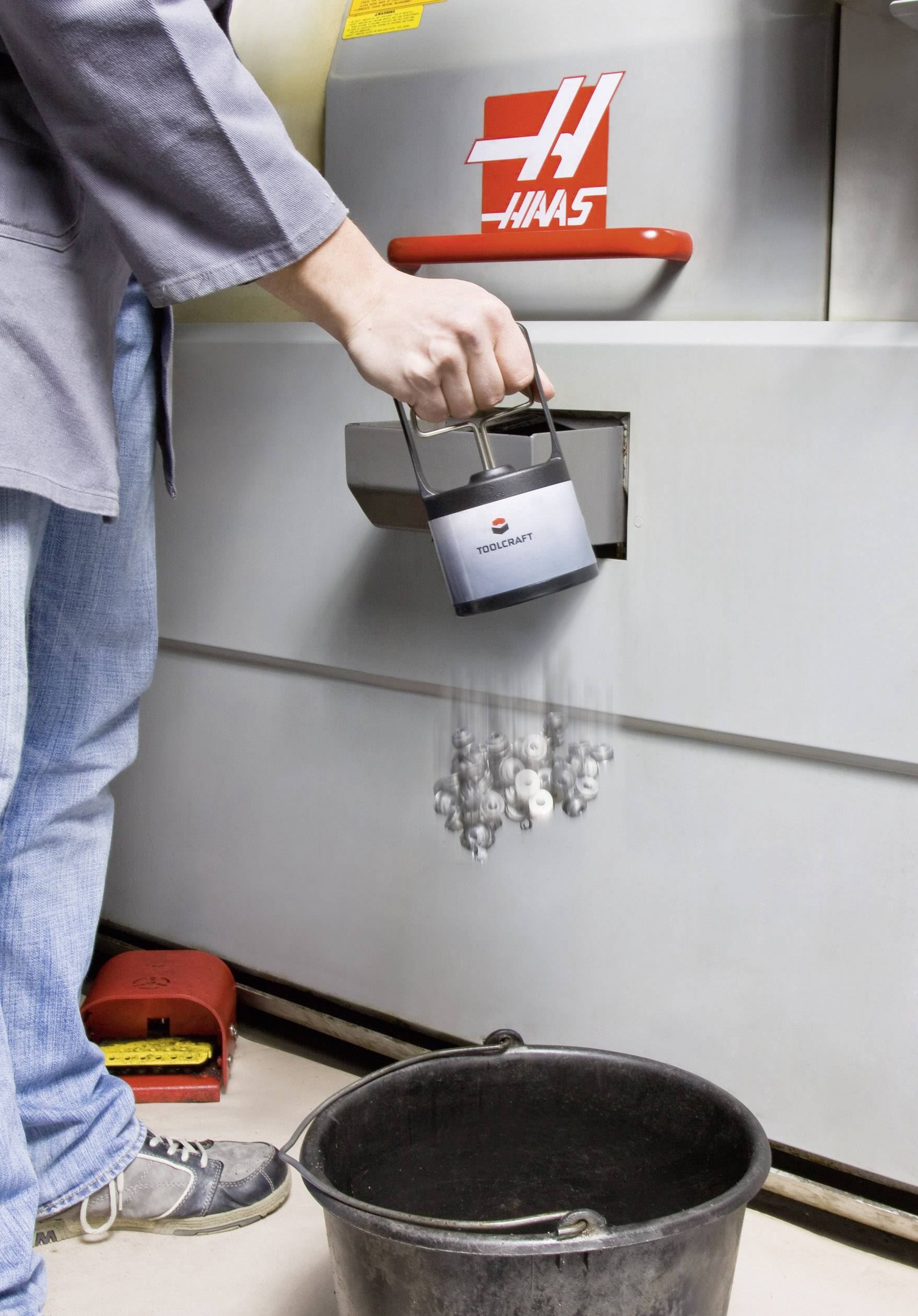 A person empties a jug with metal parts into a black bucket in front of an industrial machine, marked with a red 'Haas' logo.