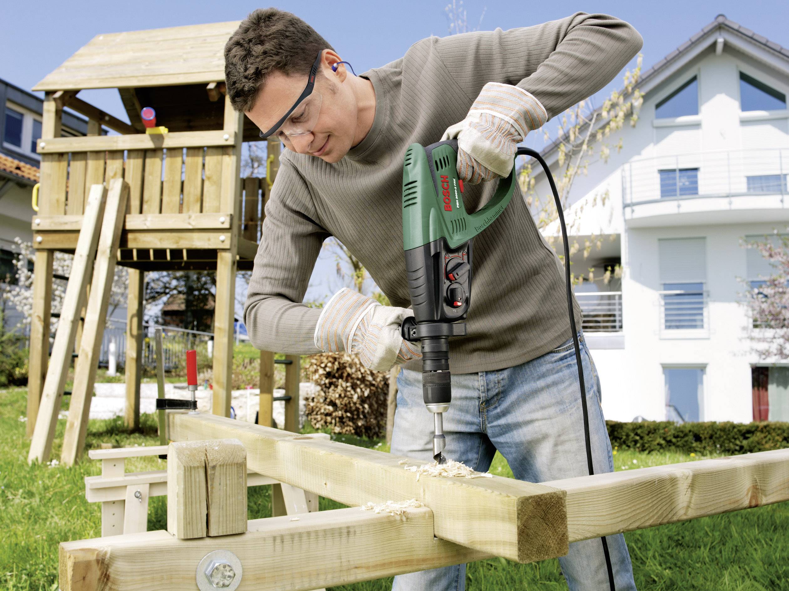 A man is drilling a hole in wooden beams outside in a garden, with a playhouse and a residential house in the background, using a green drill.