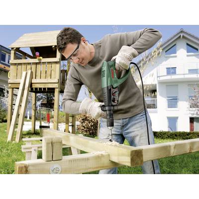 A man is drilling a hole in wooden beams outside in a garden, with a playhouse and a residential house in the background, using a green drill.