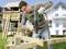 A man is drilling a hole in wooden beams outside in a garden, with a playhouse and a residential house in the background, using a green drill.