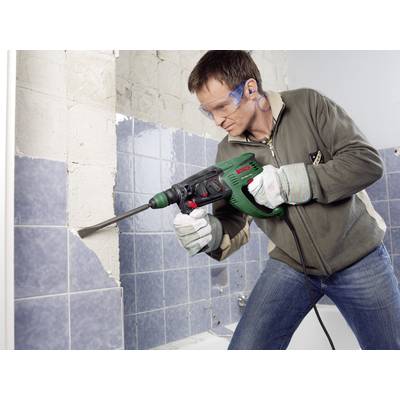 A man wearing glasses and ear protection is using a drill hammer to remove tiles from a wall.