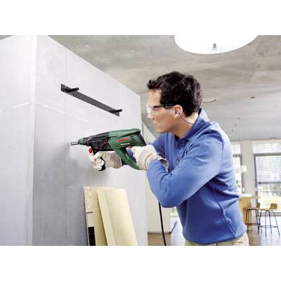 A man is drilling into a grey wall in a modern interior using a green drill. He is wearing safety glasses and ear protection.
