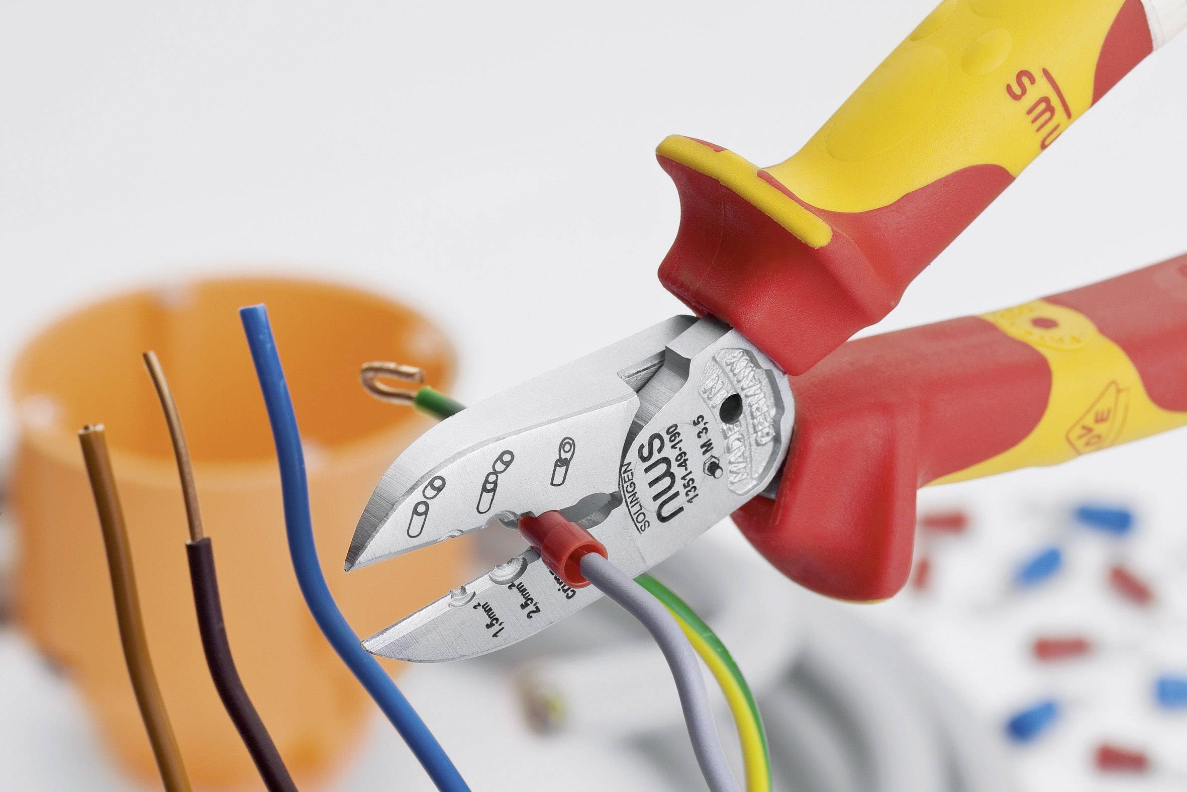 A red and yellow pair of pliers cuts through insulated wires. In the background, an orange container and a tangle of cables are visible.