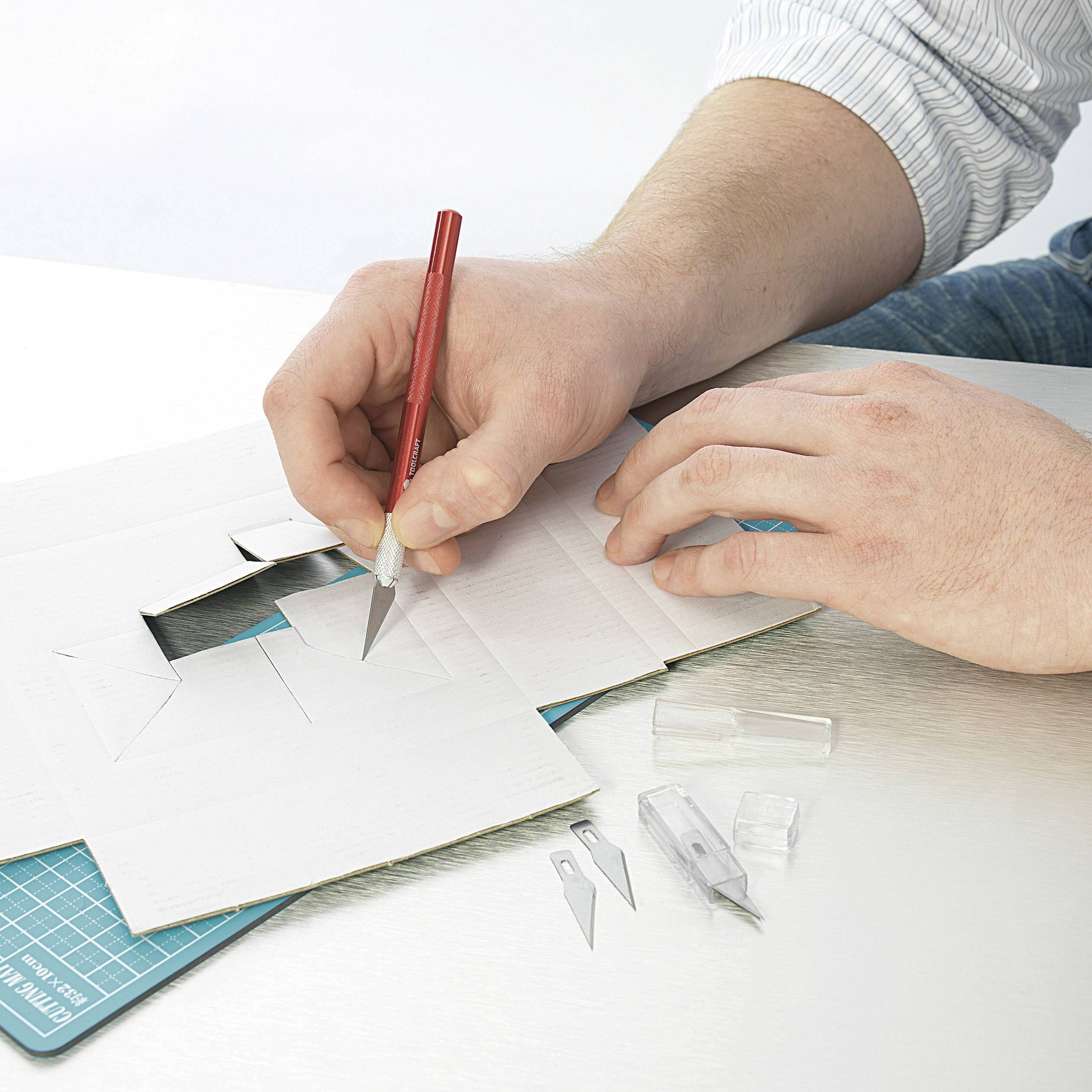 A person is using a craft knife to make precise cuts on white card. Several blades and a cutting mat are visible.