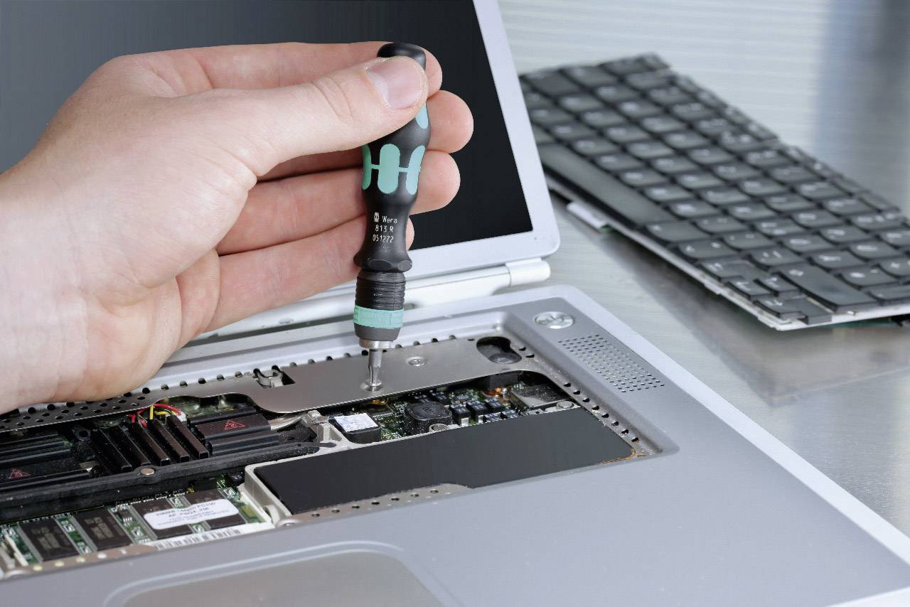 A hand is repairing an open laptop with a screwdriver. The interior of the laptop is visible. A keyboard lies in the background.