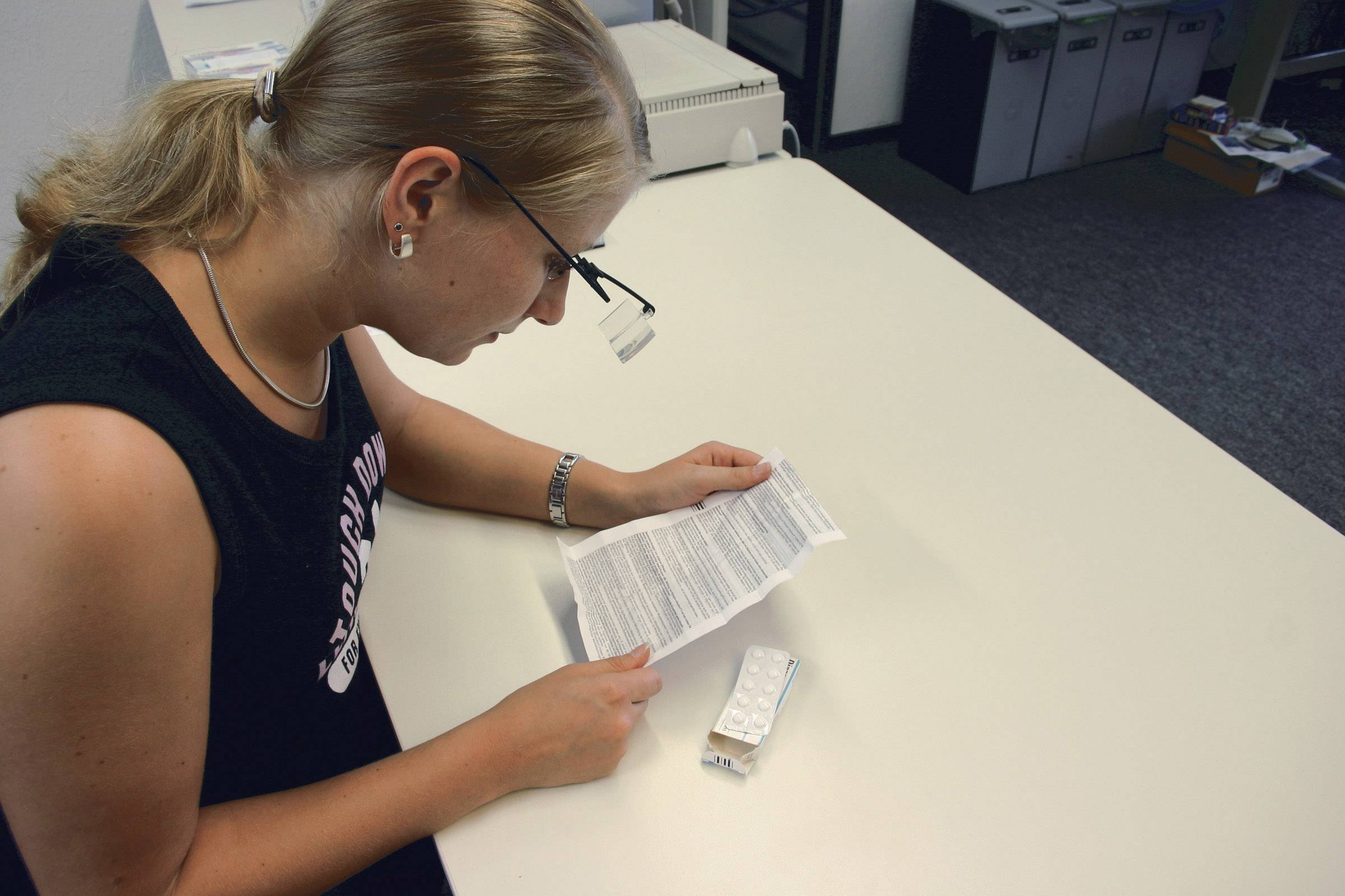A person is reading the patient information leaflet of a medication attentively, which is lying on the table. They are wearing glasses and sitting in an office.