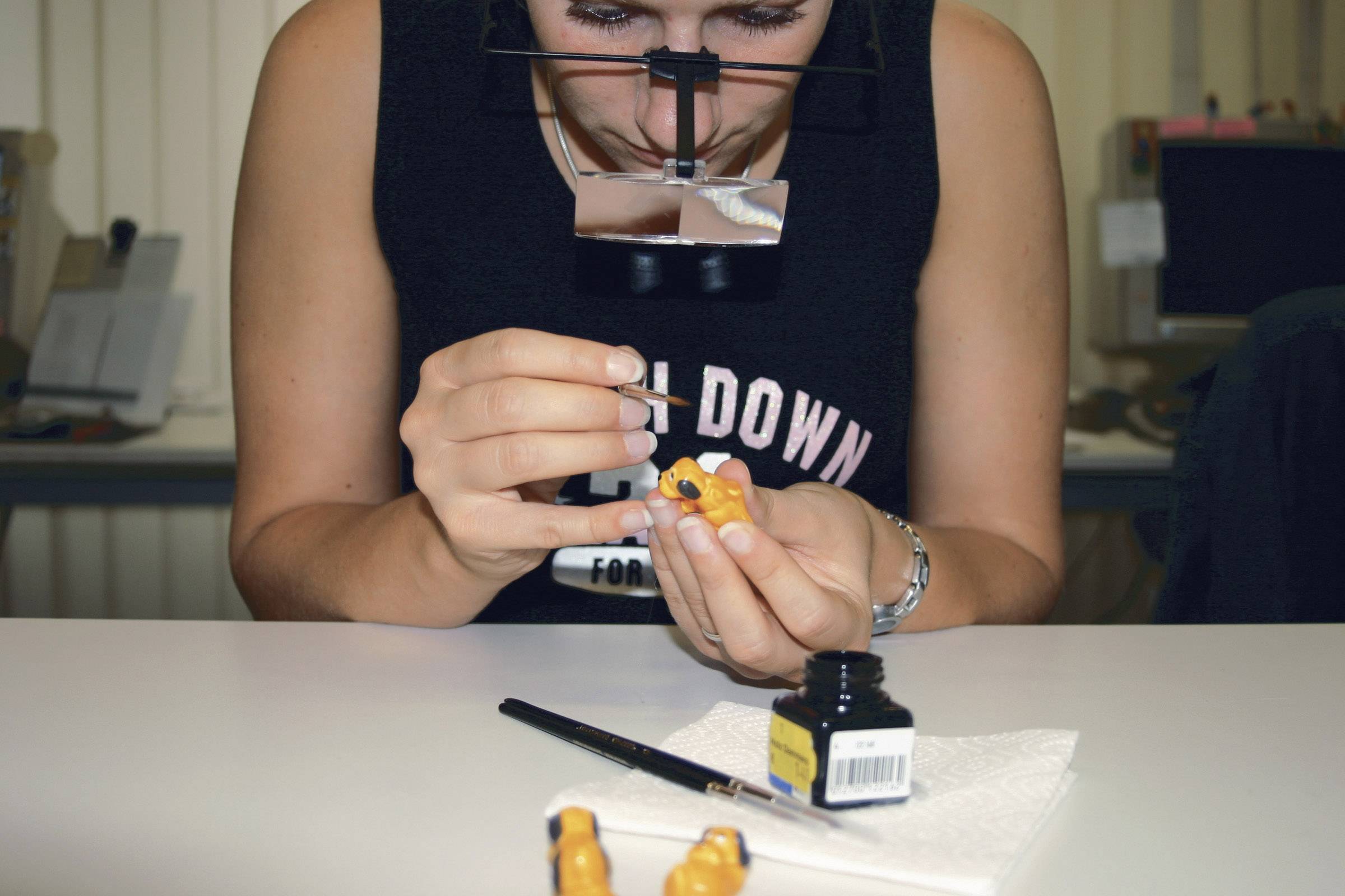 A person carefully paints small figures with a magnifying glass hanging around their neck. An inkwell and paintbrush are lying on the table.