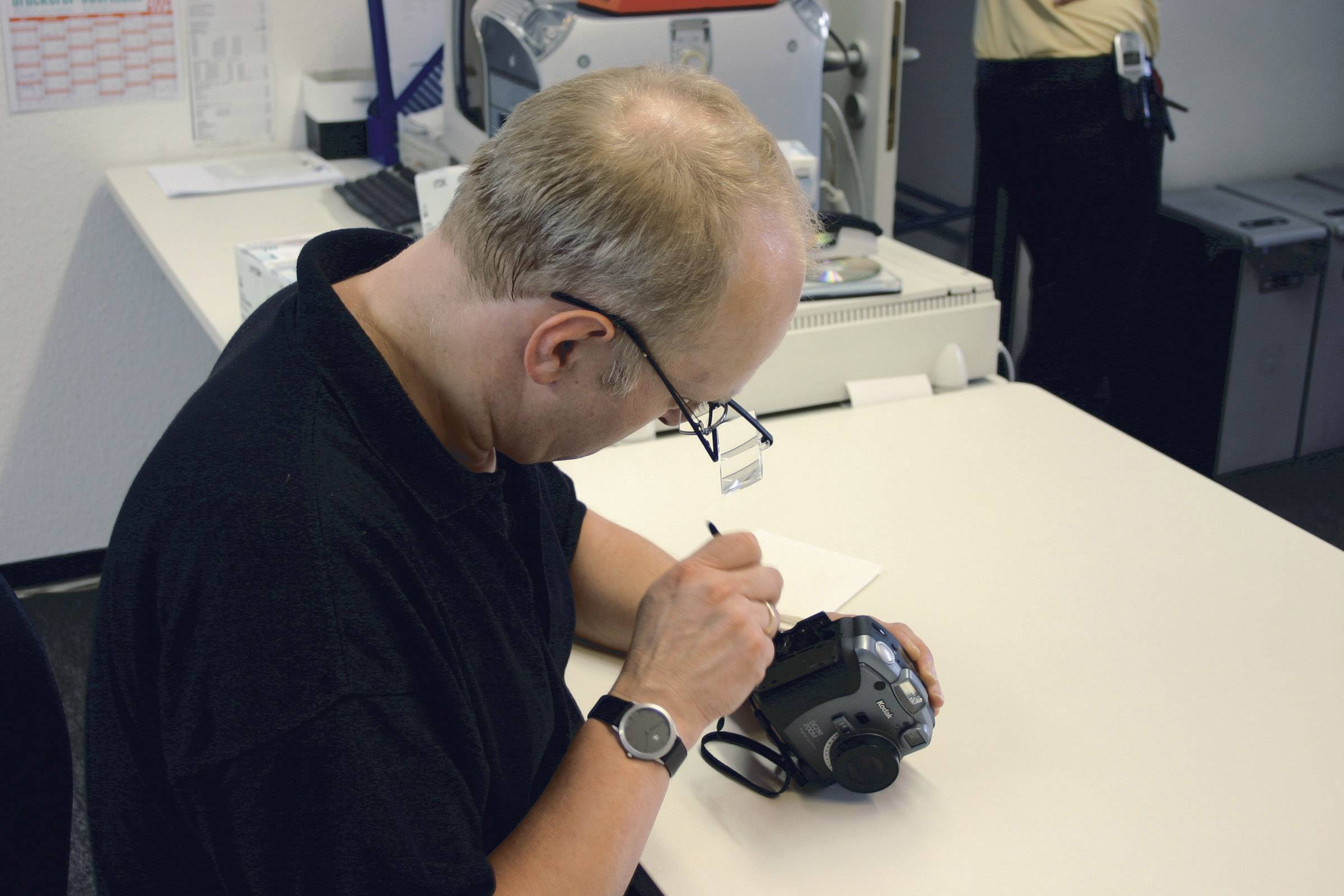A man is sitting at a table and working on a camera with a pen. Office items can be seen in the background.