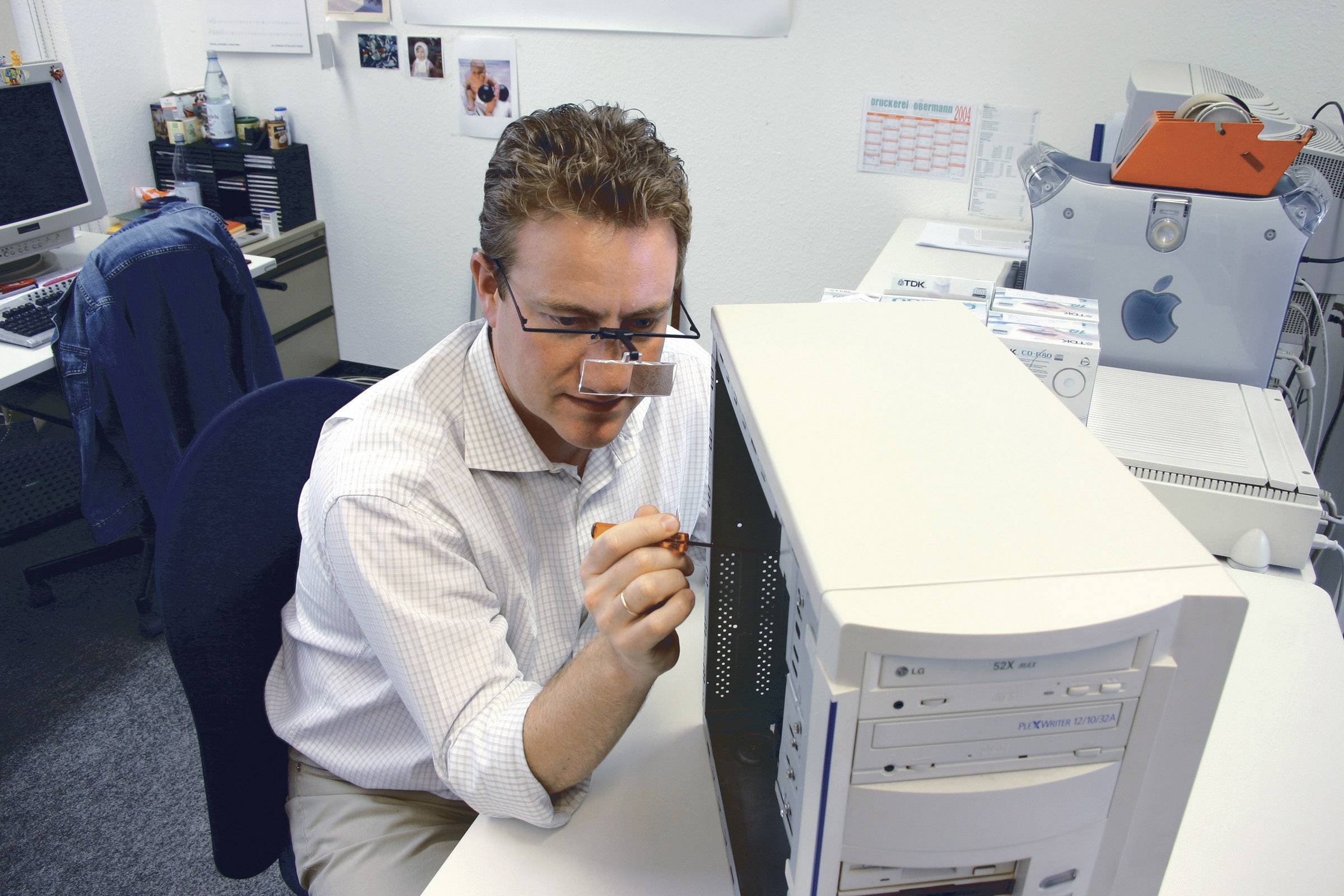 A man is checking the cable connections in an open computer case in an office with multiple computers in the background.