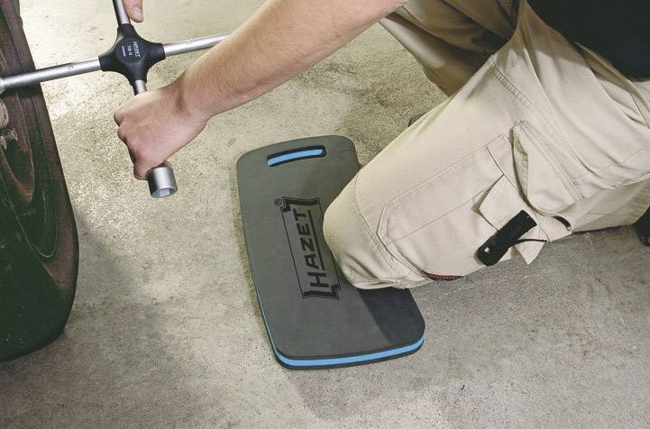 A person is kneeling on a 'Hazet' knee pad whilst using a tool to maintain or repair a vehicle wheel.