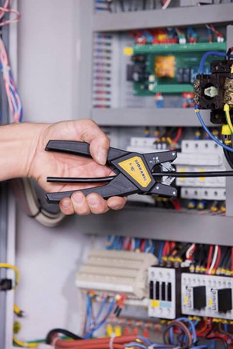 A hand holds a crimping tool in front of an open electrical cabinet with numerous cables and electrical components.