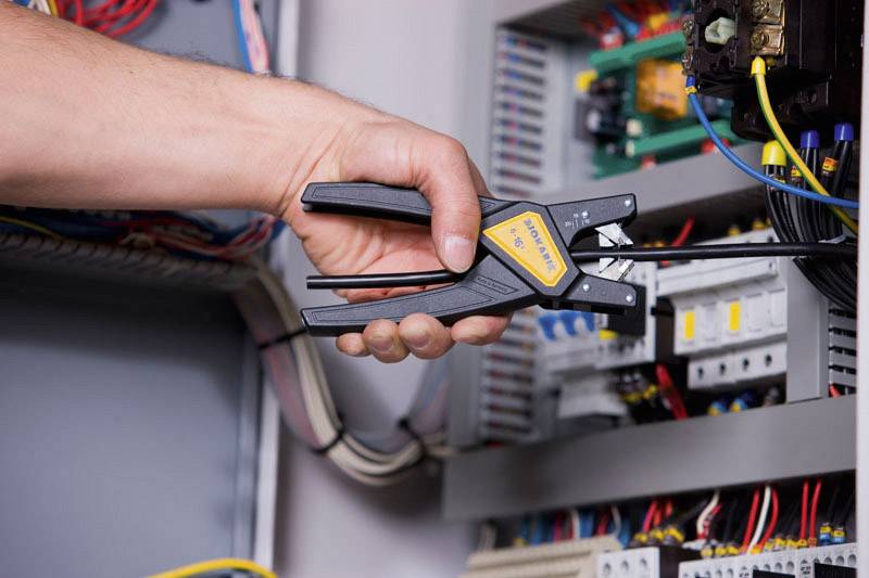 A hand is holding a tool and working on an open electrical cabinet with cables and electrical components inside.