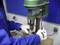 A person in blue workwear is using a pillar drill to machine a piece of metal. They are wearing white work gloves.