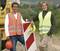 Two people in high-visibility vests are standing at a construction site, leaning against a road sign.