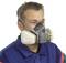 A man is wearing a respirator with filter cartridges. He has grey hair and is wearing blue protective clothing. The mask covers his mouth and nose.