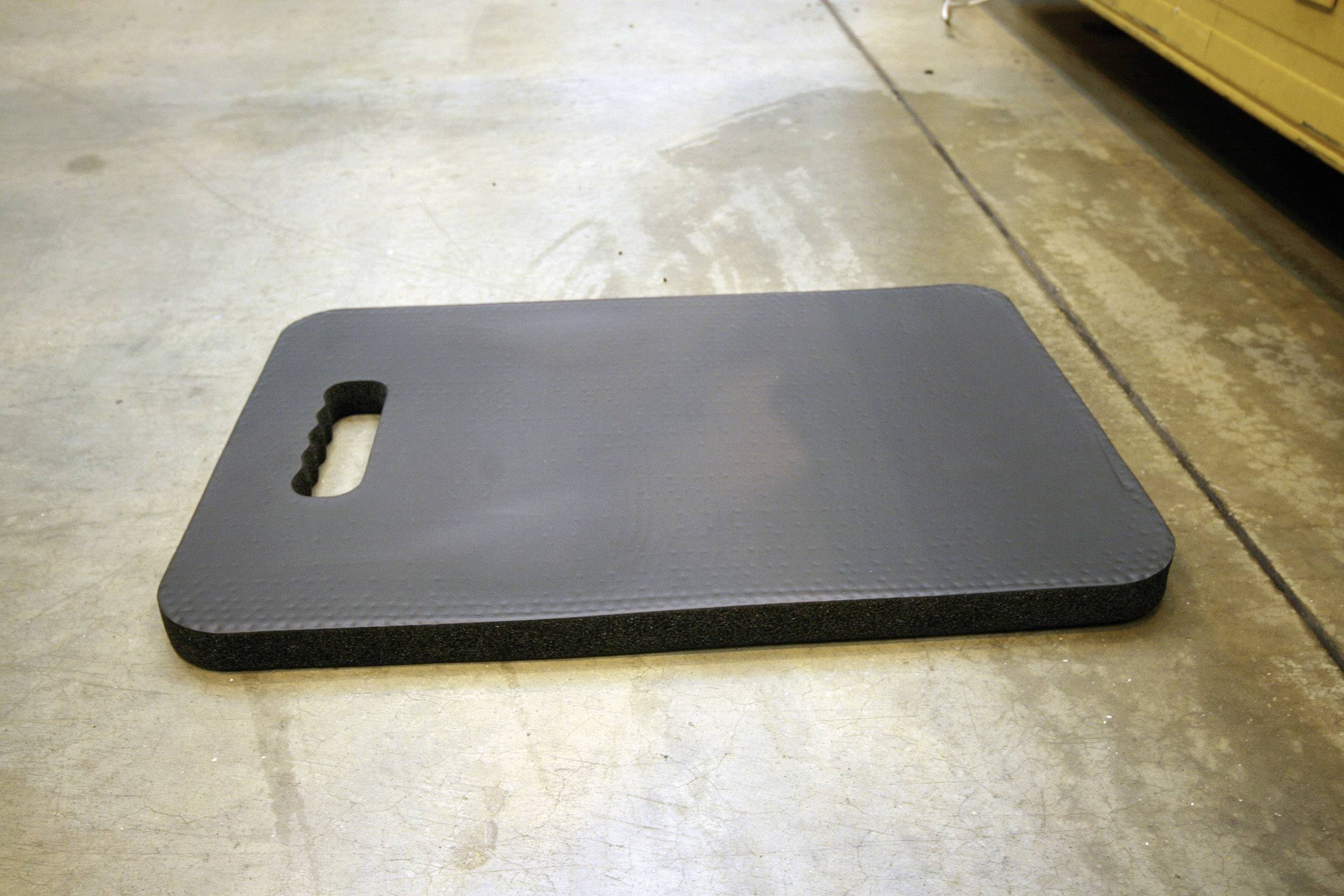 A rectangular, dark grey foam cushion with a handle lies on a concrete floor. It is used to provide knee support.