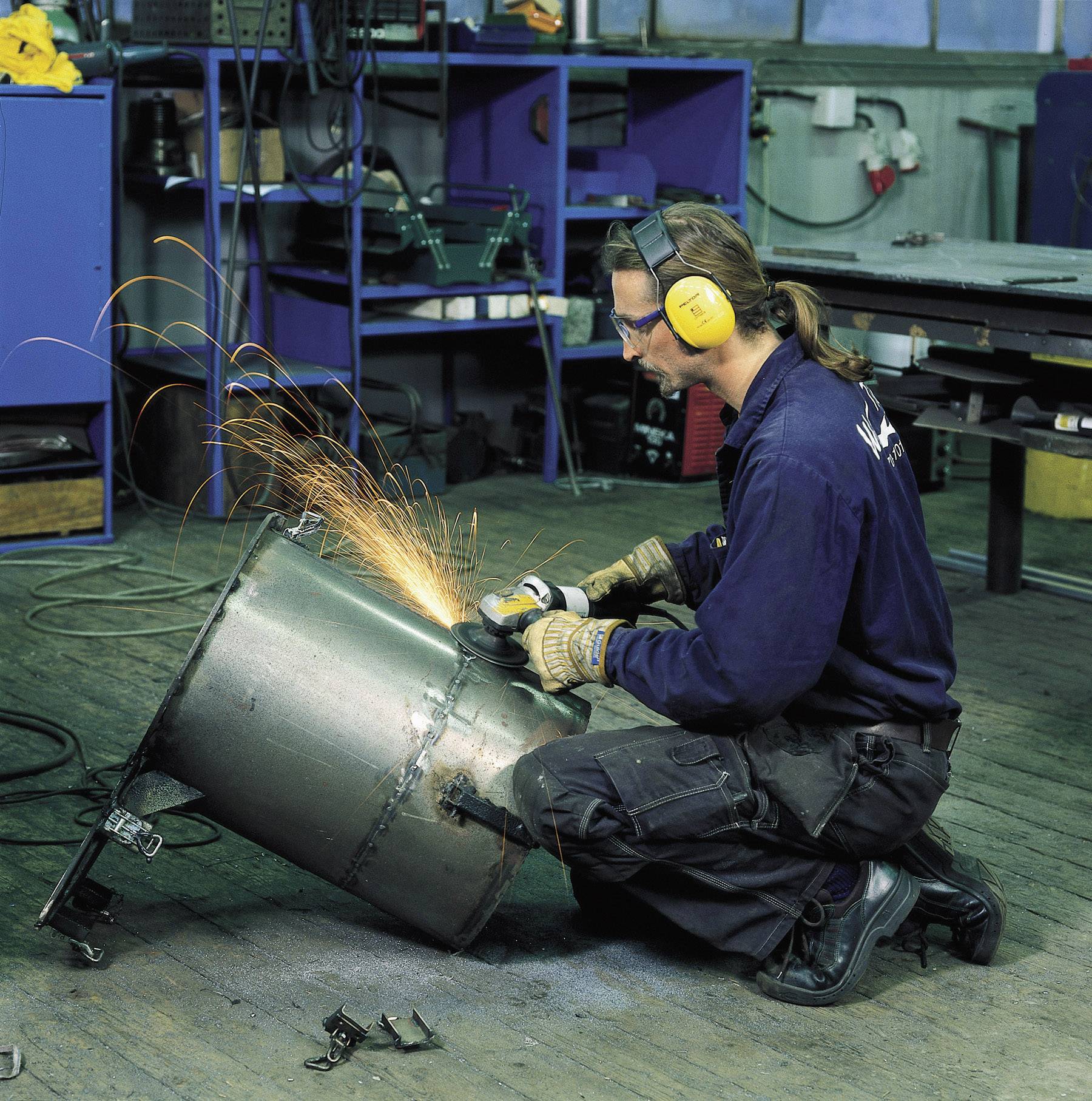 A person wearing safety glasses and ear defenders is welding, sparks flying, on a large metal cylinder in a workshop.