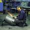 A person wearing safety glasses and ear defenders is welding, sparks flying, on a large metal cylinder in a workshop.