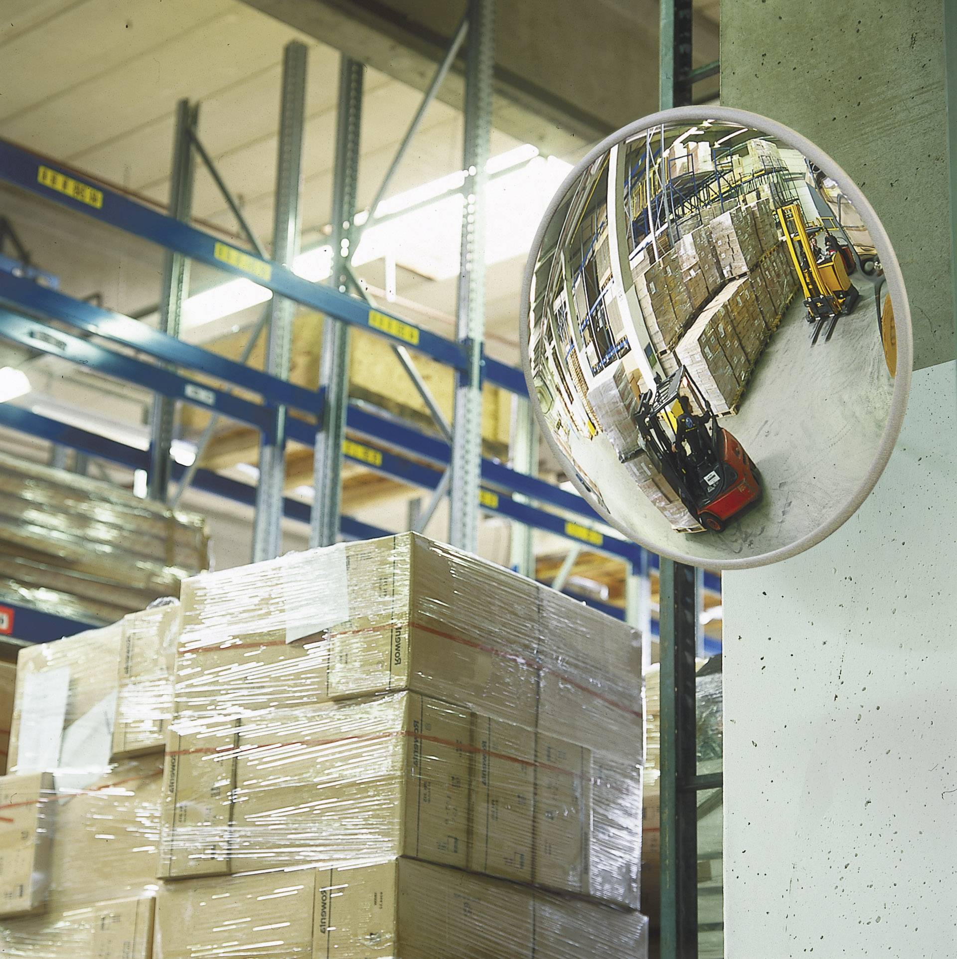 A storage room with stacked boxes on pallets. A convex mirror reflects a man on a forklift driving through the space.